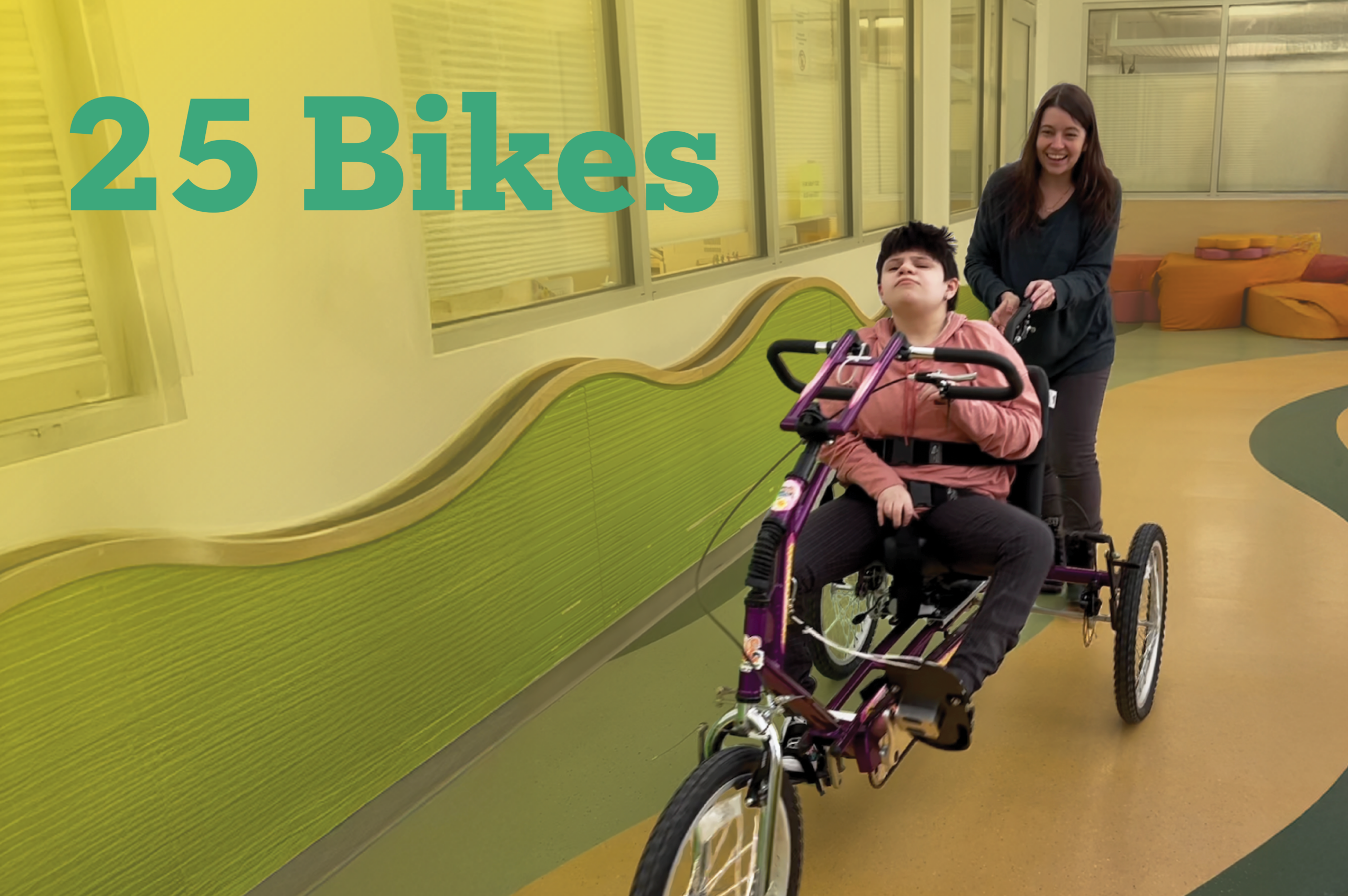 A child riding an adapted tricycle indoors, with another person walking behind and assisting. The hallway has green and yellow walls, and large text on the image reads “25 Bikes.”